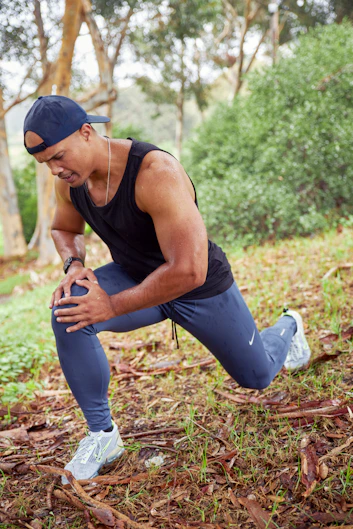 a man in a black tank top and blue pants squatting in the woods
