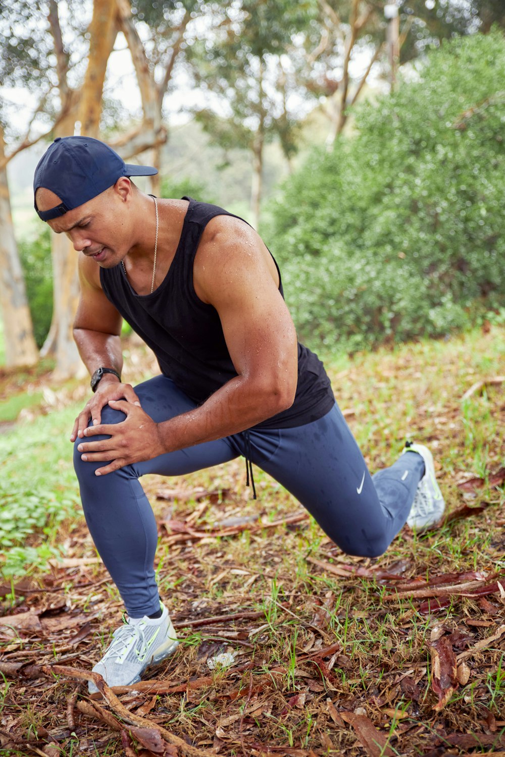 a man in a black tank top and blue pants squatting in the woods