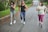 Three people are jogging on a wet road surrounded by greenery. The person on the left is wearing a gray top and light gray leggings, the person in the middle is wearing a black tank top and blue leggings, and the person on the right is wearing a light green top and purple leggings. They appear to be engaged in conversation or enjoying each other's company.