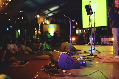 A dimly lit indoor stage features microphones and audio equipment on the floor. People are sitting casually around the stage area, creating a relaxed and intimate atmosphere. In the background, a bright screen displays text and provides the primary source of light in the room.