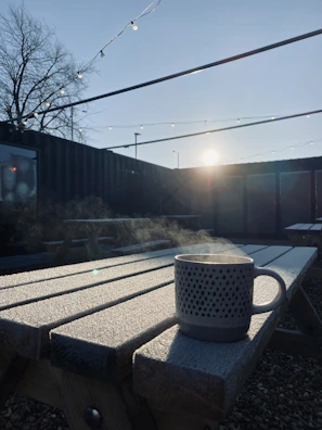 A steaming cup of coffee resting on a rustic wooden picnic table overlooking a misty trail.