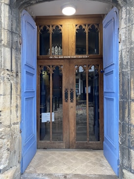 An ornate wooden door with Gothic-style arch decorations and glass panels is flanked by open blue shutters. Above the door, an inscription reads 'BE STILL AND KNOW THAT I AM GOD'. The stone frame and tiled entryway add to the historical architectural style.