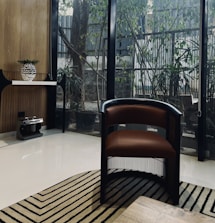 A modern interior space featuring a brown and black chair with a sleek design, placed on a patterned rug. The room has a large glass wall with a view of outdoor greenery and a wooden tabletop surface. A console table is positioned to the left, holding a decorative vase with foliage. The floor is polished and light-colored, enhancing the room's contemporary aesthetic.