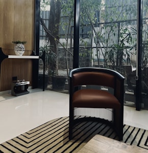 A modern interior space featuring a brown and black chair with a sleek design, placed on a patterned rug. The room has a large glass wall with a view of outdoor greenery and a wooden tabletop surface. A console table is positioned to the left, holding a decorative vase with foliage. The floor is polished and light-colored, enhancing the room's contemporary aesthetic.