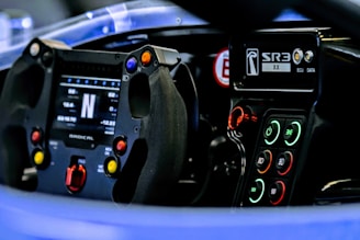 The image depicts the interior of a race car, focusing on the steering wheel and dashboard. The steering wheel is equipped with multiple colored buttons, and there is a digital display showing various metrics. The dashboard features a series of illuminated controls with indicators for systems like ignition, lights, and more.