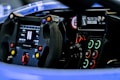 The image depicts the interior of a race car, focusing on the steering wheel and dashboard. The steering wheel is equipped with multiple colored buttons, and there is a digital display showing various metrics. The dashboard features a series of illuminated controls with indicators for systems like ignition, lights, and more.