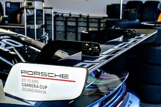A detailed view of a sports car's rear spoiler with the inscription 'Porsche 20 Years Carrera Cup Scandinavia'. Multiple tires are stacked in the background, suggesting a garage or pit environment.