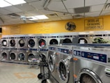 A laundromat with multiple rows of front-loading washing machines and dryers. Some machines have clothes inside, and a laundry cart is present. The walls have humorous signage, including 'Laundry today or naked tomorrow!' and 'Dryers are for washing customers only.' The flooring is light-colored tile, and there's a fan mounted on the wall.