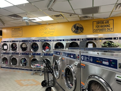 A laundromat with multiple rows of front-loading washing machines and dryers. Some machines have clothes inside, and a laundry cart is present. The walls have humorous signage, including 'Laundry today or naked tomorrow!' and 'Dryers are for washing customers only.' The flooring is light-colored tile, and there's a fan mounted on the wall.
