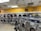 A laundromat with multiple rows of front-loading washing machines and dryers. Some machines have clothes inside, and a laundry cart is present. The walls have humorous signage, including 'Laundry today or naked tomorrow!' and 'Dryers are for washing customers only.' The flooring is light-colored tile, and there's a fan mounted on the wall.