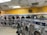 A laundromat with multiple rows of front-loading washing machines and dryers. Some machines have clothes inside, and a laundry cart is present. The walls have humorous signage, including 'Laundry today or naked tomorrow!' and 'Dryers are for washing customers only.' The flooring is light-colored tile, and there's a fan mounted on the wall.