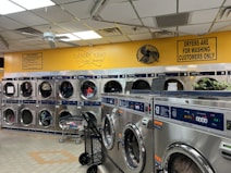 A laundromat with multiple rows of front-loading washing machines and dryers. Some machines have clothes inside, and a laundry cart is present. The walls have humorous signage, including 'Laundry today or naked tomorrow!' and 'Dryers are for washing customers only.' The flooring is light-colored tile, and there's a fan mounted on the wall.
