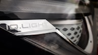 Close-up of a car headlight with a metallic surface and text that reads 'IQ.LIGHT'. The design is sleek, with a combination of smooth and textured elements. The image has a reflective and shiny appearance.