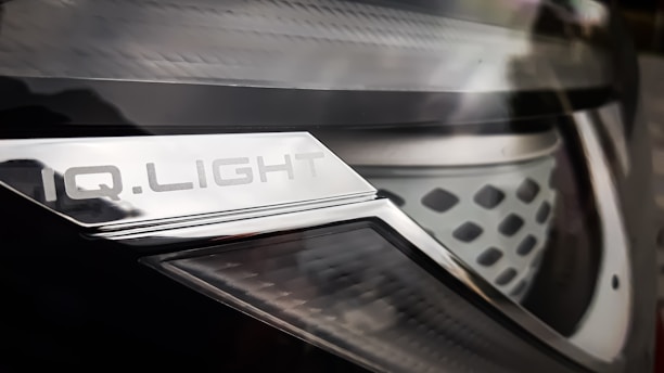 Close-up of a car headlight with a metallic surface and text that reads 'IQ.LIGHT'. The design is sleek, with a combination of smooth and textured elements. The image has a reflective and shiny appearance.
