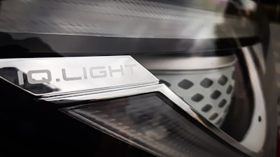 Close-up of a car headlight with a metallic surface and text that reads 'IQ.LIGHT'. The design is sleek, with a combination of smooth and textured elements. The image has a reflective and shiny appearance.