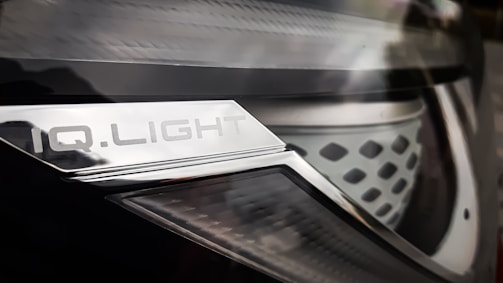 Close-up of a car headlight with a metallic surface and text that reads 'IQ.LIGHT'. The design is sleek, with a combination of smooth and textured elements. The image has a reflective and shiny appearance.