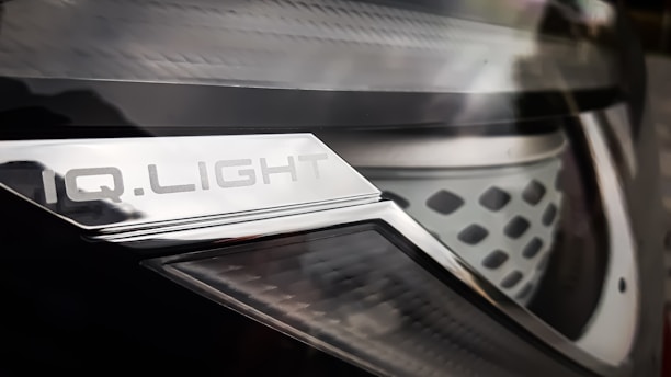 Close-up of a car headlight with a metallic surface and text that reads 'IQ.LIGHT'. The design is sleek, with a combination of smooth and textured elements. The image has a reflective and shiny appearance.