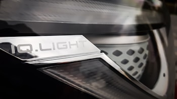 Close-up of a car headlight with a metallic surface and text that reads 'IQ.LIGHT'. The design is sleek, with a combination of smooth and textured elements. The image has a reflective and shiny appearance.