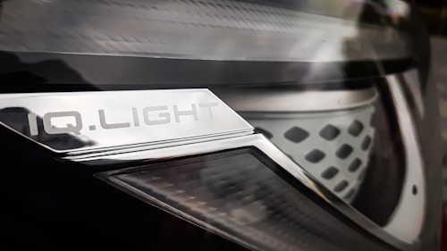Close-up of a car headlight with a metallic surface and text that reads 'IQ.LIGHT'. The design is sleek, with a combination of smooth and textured elements. The image has a reflective and shiny appearance.
