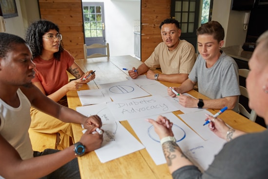A group of people sit around a wooden table engaged in a discussion, with sheets of paper in front of them marked with diagrams and the words 'Diabetes Advocacy'. There is a bright, natural light coming from a nearby window, and everyone appears focused on sharing ideas, as some take notes with colored markers.