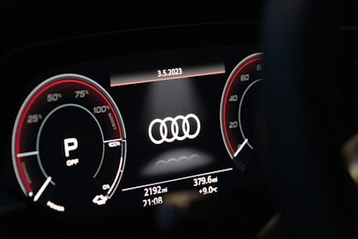 A close-up of a digital car dashboard featuring the Audi logo at the center. Circular gauges on either side display information such as speed and engine RPM, with red accents and various numerical indicators. The date, odometer reading, mileage, time, and temperature are shown on the bottom of the display.