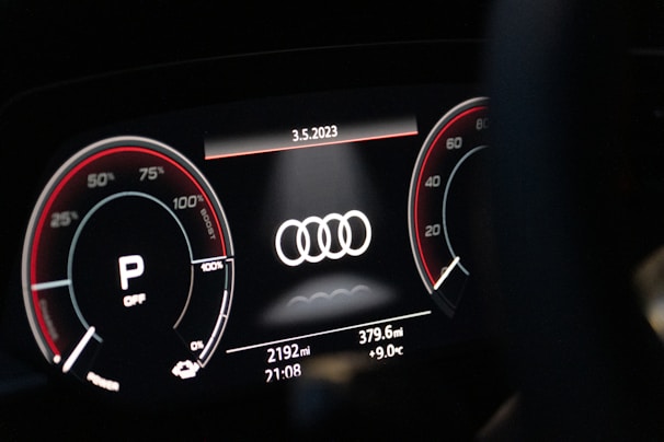 A close-up of a digital car dashboard featuring the Audi logo at the center. Circular gauges on either side display information such as speed and engine RPM, with red accents and various numerical indicators. The date, odometer reading, mileage, time, and temperature are shown on the bottom of the display.