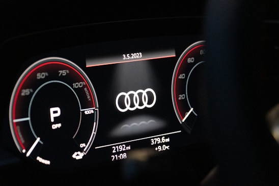 A close-up of a digital car dashboard featuring the Audi logo at the center. Circular gauges on either side display information such as speed and engine RPM, with red accents and various numerical indicators. The date, odometer reading, mileage, time, and temperature are shown on the bottom of the display.