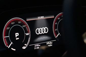 A close-up of a digital car dashboard featuring the Audi logo at the center. Circular gauges on either side display information such as speed and engine RPM, with red accents and various numerical indicators. The date, odometer reading, mileage, time, and temperature are shown on the bottom of the display.