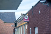 A brick building with a sign on the side that says 'payzone'. The sign features a logo above the text. Security cameras are mounted on the building, and a metal structure is attached to the roof. The background includes part of another brick building.