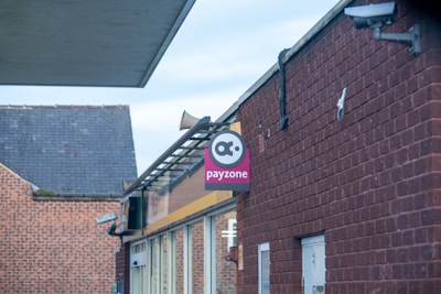 A brick building with a sign on the side that says 'payzone'. The sign features a logo above the text. Security cameras are mounted on the building, and a metal structure is attached to the roof. The background includes part of another brick building.