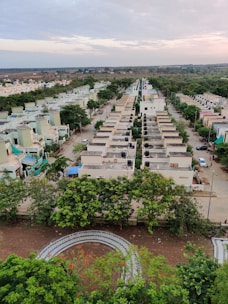 Aerial view of a residential neighborhood with uniform houses lined up along the streets. The trees are lush and green, creating a vibrant contrast with the muted colors of the houses. The sky is partly cloudy, indicating a calm and overcast day. In the foreground, a curved path surrounded by greenery is visible.