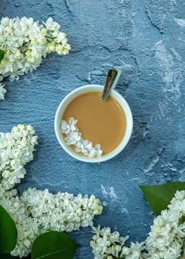 A cup filled with coffee or tea, featuring a spoon, is surrounded by delicate white flowers on a textured blue surface. The flowers are carefully placed around the cup, creating a serene and aesthetically pleasing arrangement.