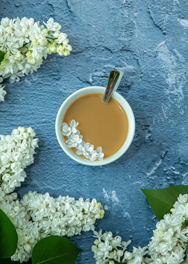 A cup filled with coffee or tea, featuring a spoon, is surrounded by delicate white flowers on a textured blue surface. The flowers are carefully placed around the cup, creating a serene and aesthetically pleasing arrangement.