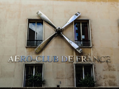A building facade features two windows with reflective glass. Between the windows, there is a large propeller mounted on the wall. Below the propeller, the words 'AERO-CLUB DE FRANCE' are prominently displayed in metallic letters.