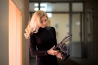 A person with long blonde hair wearing a black outfit is holding a professional camera with attached equipment, including a screen and a microphone. The scene is set in a softly lit indoor corridor, with several blurred rectangular lights in the background.