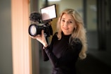 A person with long blonde hair is holding a professional camera equipped with a furry microphone and a large lens. The person is wearing a black turtleneck sweater and standing indoors, with a softly blurred background.