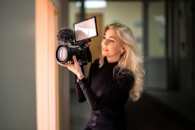 A person with long blonde hair is holding a professional camera with a large lens and an attached microphone. They seem focused and engaged in the act of recording or photographing. The setting appears to be indoors with soft ambient lighting.