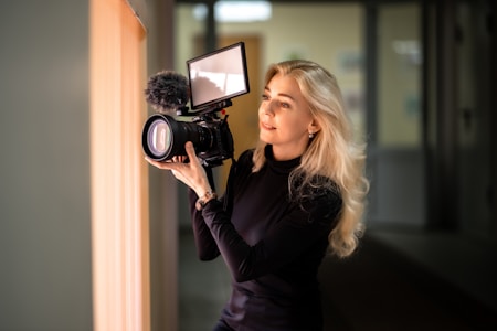 A person with long blonde hair is holding a professional camera with a large lens and an attached microphone. They seem focused and engaged in the act of recording or photographing. The setting appears to be indoors with soft ambient lighting.
