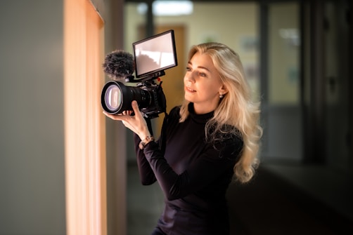 A person with long blonde hair is holding a professional camera with a large lens and an attached microphone. They seem focused and engaged in the act of recording or photographing. The setting appears to be indoors with soft ambient lighting.