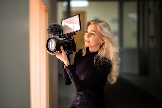A person with long blonde hair is holding a professional camera with a large lens and an attached microphone. They seem focused and engaged in the act of recording or photographing. The setting appears to be indoors with soft ambient lighting.
