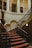 Grand staircase adorned with ornate railings and red patterned carpet. The staircase is set within an elegant and historic interior, with a large clock mounted on the wall, arches, columns, and framed portraits hanging on the walls. The lighting is warm and creates a sophisticated ambiance.