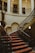 Grand staircase adorned with ornate railings and red patterned carpet. The staircase is set within an elegant and historic interior, with a large clock mounted on the wall, arches, columns, and framed portraits hanging on the walls. The lighting is warm and creates a sophisticated ambiance.