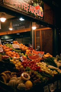 A richly stocked fruit and vegetable market stall displays a variety of fresh produce, including watermelons, oranges, tomatoes, peppers, bananas, grapes, and other colorful fruits and vegetables. The stall is lit with warm lights and has handwritten price tags on the produce.