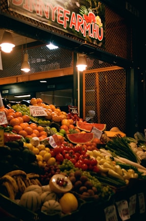 A richly stocked fruit and vegetable market stall displays a variety of fresh produce, including watermelons, oranges, tomatoes, peppers, bananas, grapes, and other colorful fruits and vegetables. The stall is lit with warm lights and has handwritten price tags on the produce.