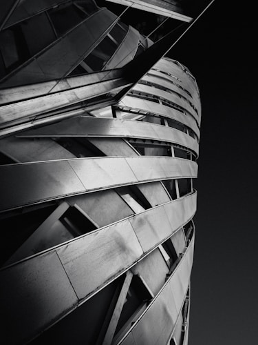 A modern architectural structure with a spiral design composed of metallic panels and angular lines. The building features a dynamic, twisted form against a stark, dark background.
