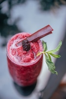 A vibrant red fruit smoothie served in a tall glass is topped with a dark berry and garnished with a sprig of fresh mint. A thin, stick-like garnish rests on top, adding a refined touch to the presentation.