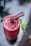 A vibrant red fruit smoothie served in a tall glass is topped with a dark berry and garnished with a sprig of fresh mint. A thin, stick-like garnish rests on top, adding a refined touch to the presentation.