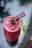 A vibrant red fruit smoothie served in a tall glass is topped with a dark berry and garnished with a sprig of fresh mint. A thin, stick-like garnish rests on top, adding a refined touch to the presentation.