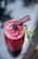 A vibrant red fruit smoothie served in a tall glass is topped with a dark berry and garnished with a sprig of fresh mint. A thin, stick-like garnish rests on top, adding a refined touch to the presentation.