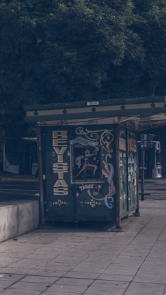 A small, decorated kiosk sits on a sidewalk, featuring colorful artwork and lettering, likely serving as a magazine or newspaper stand. It is surrounded by trees, and part of a public transport vehicle is visible in the background.